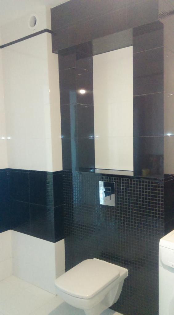 Nowoczesna łazienka z biało-czarnymi płytkami, widoczna toaleta podwieszana z chromowanym przyciskiem spłukującym i fragment pralki.