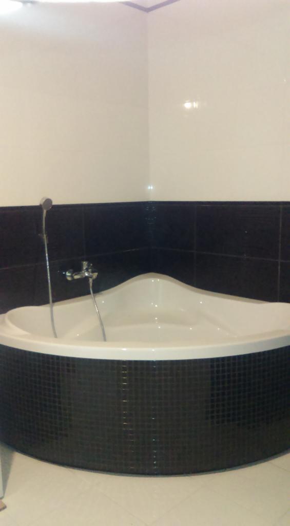 Narożna wanna obudowana czarną mozaiką w łazience z białymi płytkami na górze i jasną podłogą, widoczna bateria i słuchawka prysznicowa.