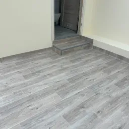 Szare panele podłogowe imitujące drewno, ułożone w jodełkę, z widocznym wejściem do łazienki z jednym stopniem i fragmentem toalety w tle.