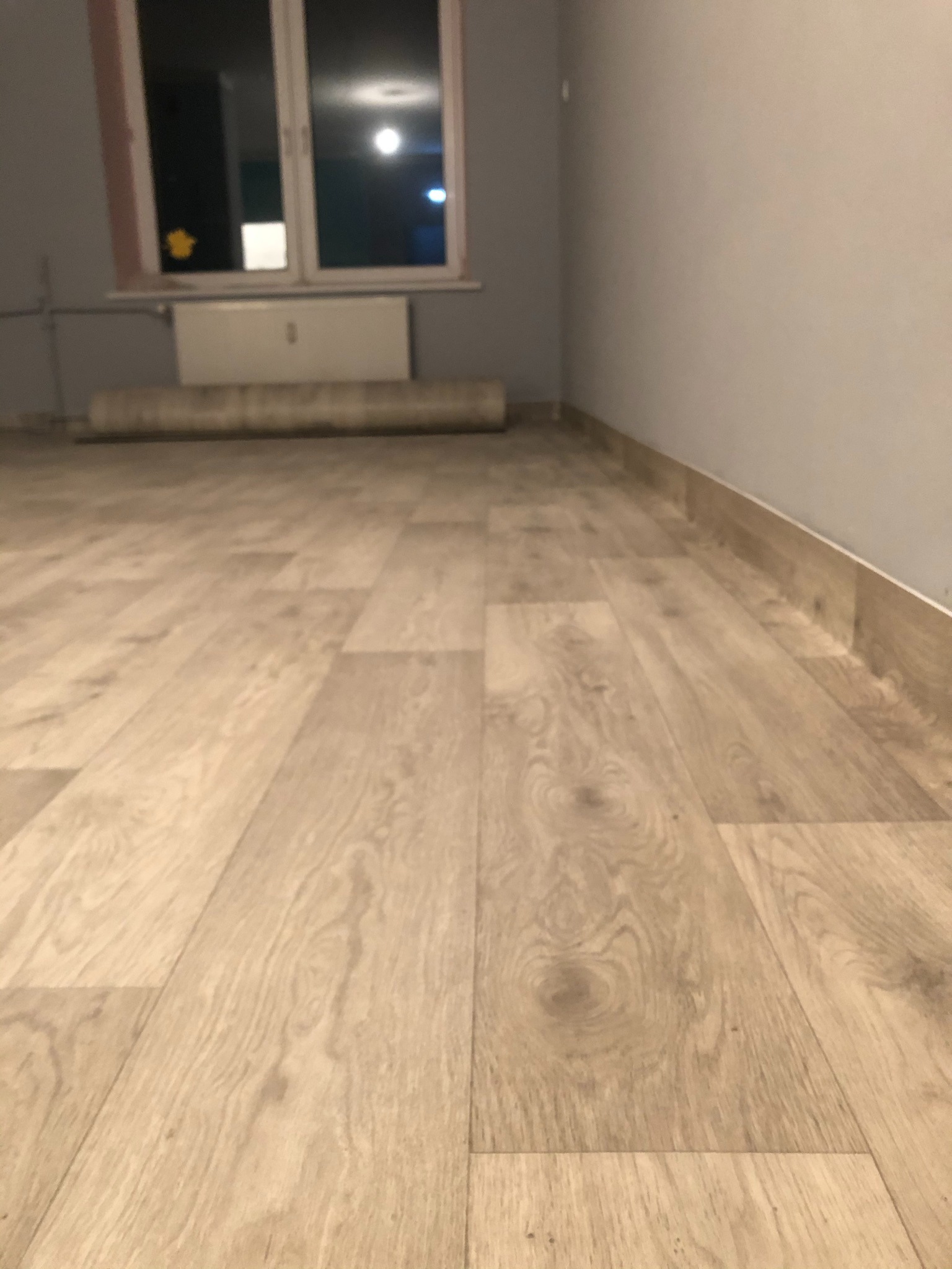 Jasne panele podłogowe imitujące drewno, ułożone w nowym pomieszczeniu z szarą ścianą i listwami przypodłogowymi, z rolką materiału pod oknem.