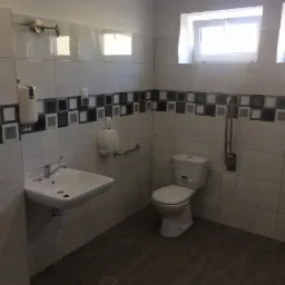 Wykończona łazienka z białymi płytkami z dekoracyjnym pasem z szarych i czarnych kwadratów, umywalka z baterią, toaleta i uchwyty dla niepełnosprawnych.