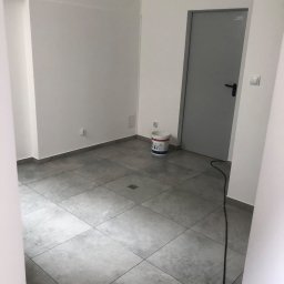 Kamil - Pomalowane na biało ściany biura z szarymi płytkami podłogowymi i szarymi drzwiami, wiadro z farbą stoi przy drzwiach.