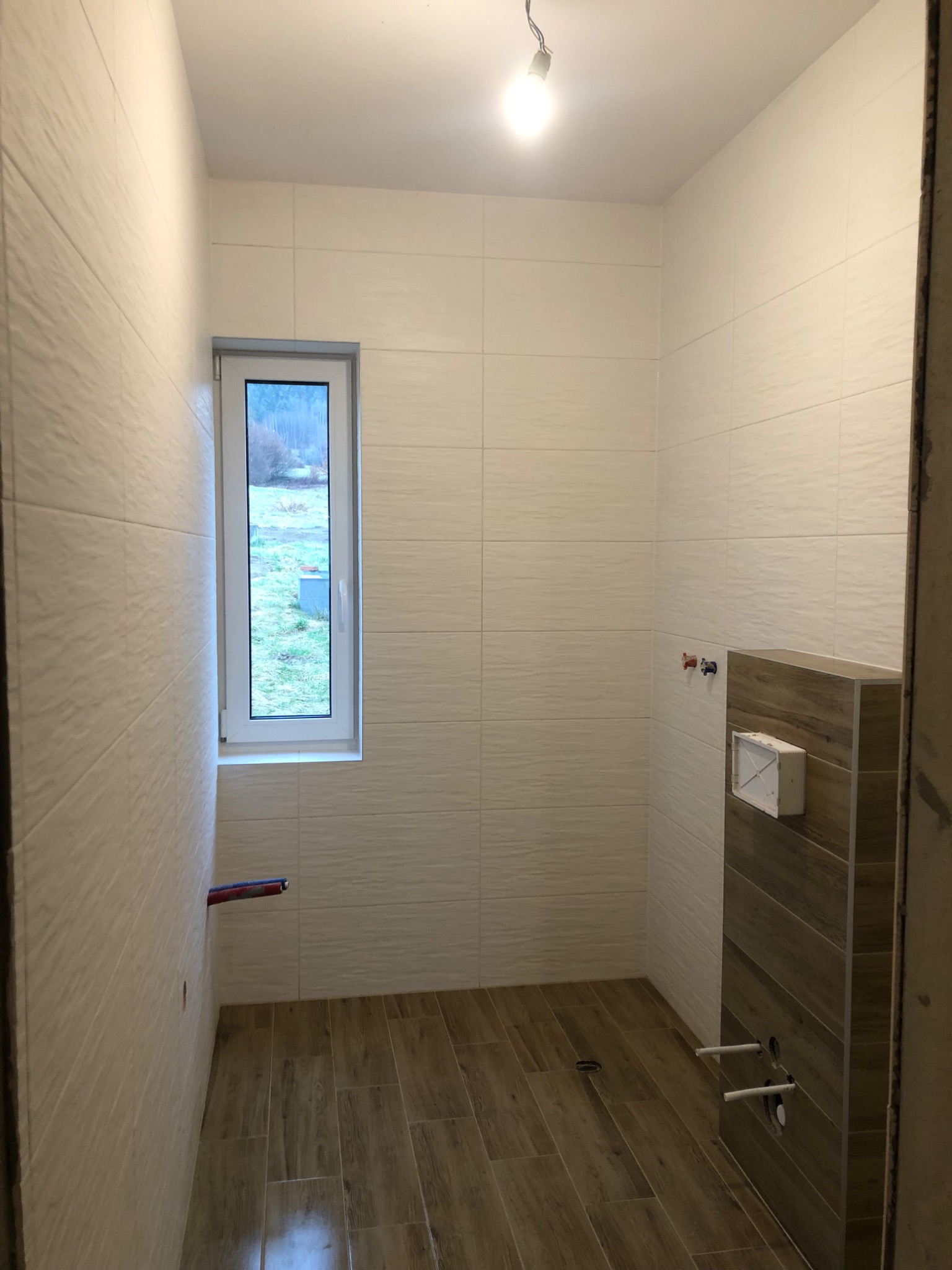 Biała łazienka z drewnopodobną podłogą i zabudową WC, widoczne wystające rury instalacji wodnej i okno z widokiem na zewnątrz.