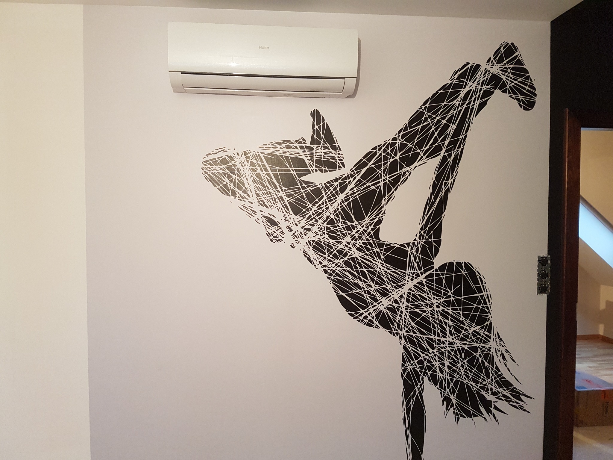 Biała klimatyzacja Haier zamontowana na ścianie z czarną, abstrakcyjną naklejką przedstawiającą tancerkę.