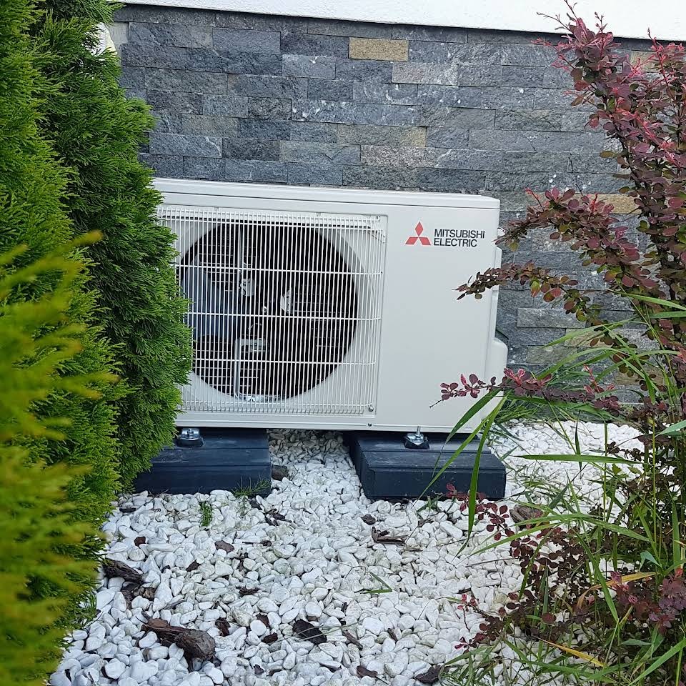 Zewnętrzna jednostka klimatyzacji Mitsubishi Electric umieszczona na betonowych podstawach, otoczona białym kamieniem i roślinnością, na tle ściany z szarego kamienia.