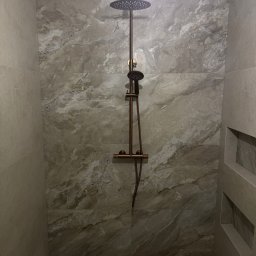 Fachowiec - Wykończona kabina prysznicowa z marmurowymi płytkami i miedzianą armaturą. Widoczne wnęki w ścianie i odpływ liniowy. Minimalistyczny design.