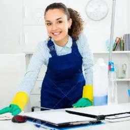 Uśmiechnięta kobieta w niebieskim kombinezonie i żółto-zielonych rękawiczkach przeciera białą ściereczką biurko w biurze, na którym leżą dokumenty i akcesoria biurowe, w tle widoczny regał...