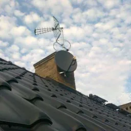 Antena telewizyjna Yagi-Uda i satelitarna zamontowane na kominie z cegły na dachu pokrytym ciemną dachówką, pochmurne niebo w tle.