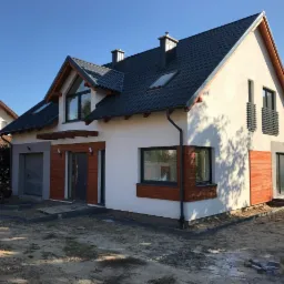 Nowoczesny dom jednorodzinny w Żukowie z elementami drewnianymi na elewacji, ciemna dachówka i jasna elewacja, widoczne prace wykończeniowe wokół budynku.