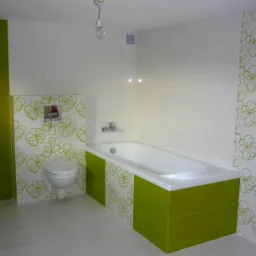 Wykończona łazienka z zielonymi akcentami: zielone płytki na ścianie i obudowie wanny, wzorzyste płytki z motywem kwiatowym na ścianie i obudowie toalety, biała wanna i podwieszana toaleta, szare...