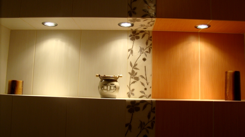 Półka w łazience z oświetleniem punktowym, dekoracyjnymi płytkami z motywem roślinnym i ceramicznym naczyniem ozdobnym.