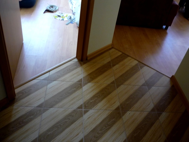 Ukośne ułożenie płytek podłogowych imitujących drewno, przechodzące w jasne panele podłogowe w progu drzwi.