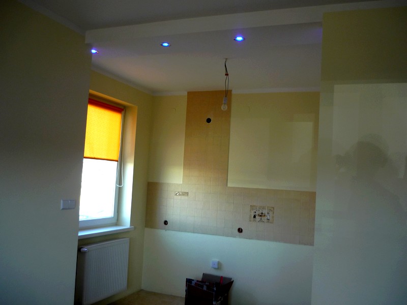 Wnętrze w trakcie prac przygotowawczych, widoczne fragmenty starej glazury, ściany przygotowane pod malowanie, okno z pomarańczową roletą, niebieskie oświetlenie punktowe w suficie.