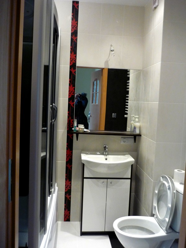 Ukończona łazienka z białą armaturą, umywalką z szafką i toaletą. Dekoracyjny pas z motywem kwiatowym na ścianie. W lustrze odbija się osoba robiąca zdjęcie.