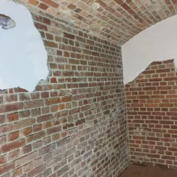 Wnętrze piwnicy z odsłoniętą cegłą na łukowym suficie i ścianach, częściowo pokrytą białym tynkiem, widoczne rury instalacyjne.