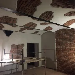 Wnętrze w trakcie prac murarskich: odsłonięta cegła na sklepieniu i ścianie, częściowo otynkowana, widoczne rusztowania i metalowe belki stropowe.