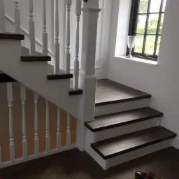 Fragment białych schodów z ciemnymi drewnianymi stopniami, białą balustradą i czarną poręczą, na podłodze z parkietu ułożonego w jodełkę leżą kapcie, w tle okno z czarną ramą i białą firanką.