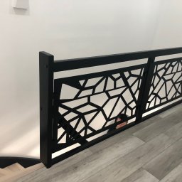 Usługowy Zakład Stolarski - Schody Drewniane - Nowoczesna czarna balustrada z geometrycznym ażurem, widok z góry na schody z drewnopodobnymi panelami, fragment osoby w tle.
