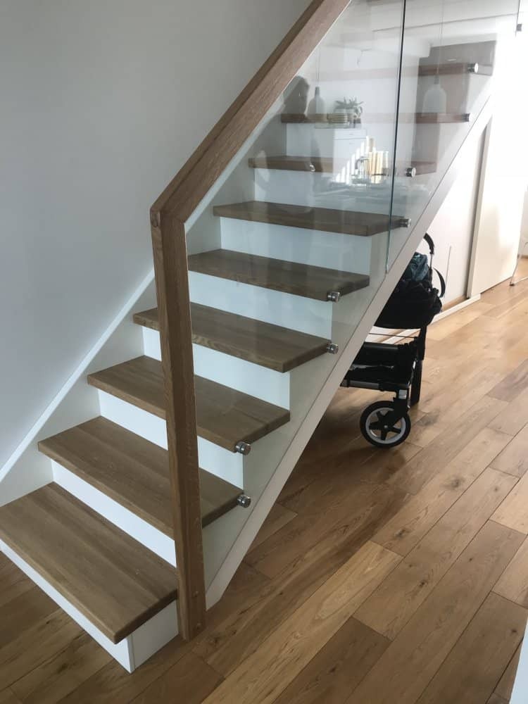 Nowoczesne schody z drewnianymi stopniami, białymi podstopnicami i szklaną balustradą mocowaną na metalowych elementach, widok z dołu na drewnianą podłogę.