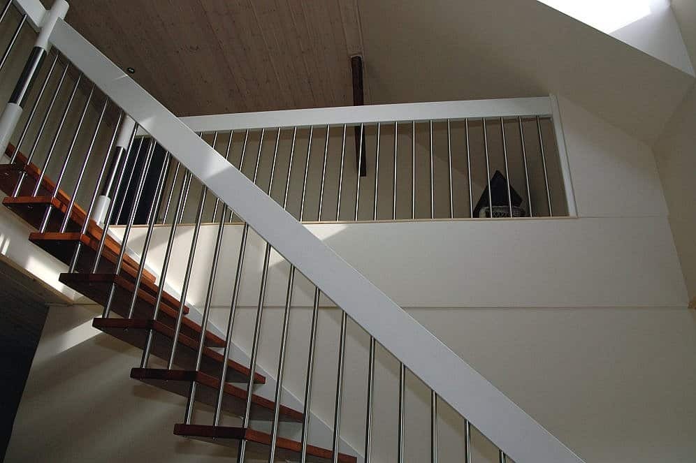 Bieg schodów z drewnianymi stopniami i balustradą z nierdzewnych prętów, widok z dołu na antresolę z podobną balustradą.