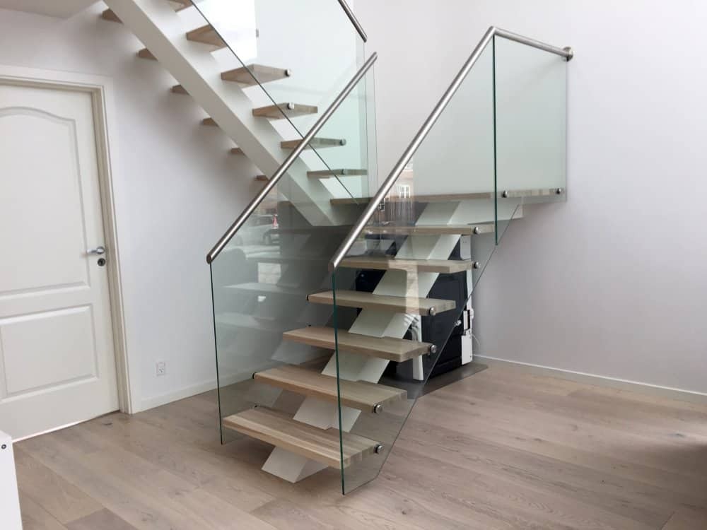 Nowoczesne schody z drewnianymi stopniami, białymi wspornikami i szklaną balustradą z metalowymi poręczami w minimalistycznym wnętrzu.