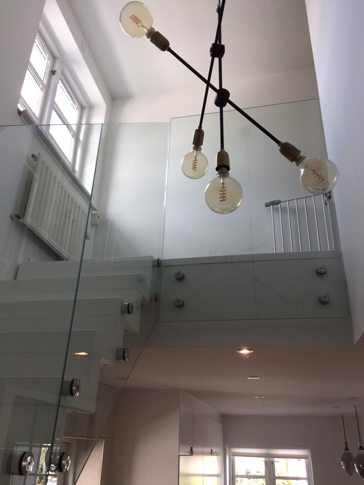 Nowoczesne białe schody z przeszkloną balustradą, oświetlone designerską lampą z żarówkami Edisona, widok z dołu na antresolę z barierką zabezpieczającą.