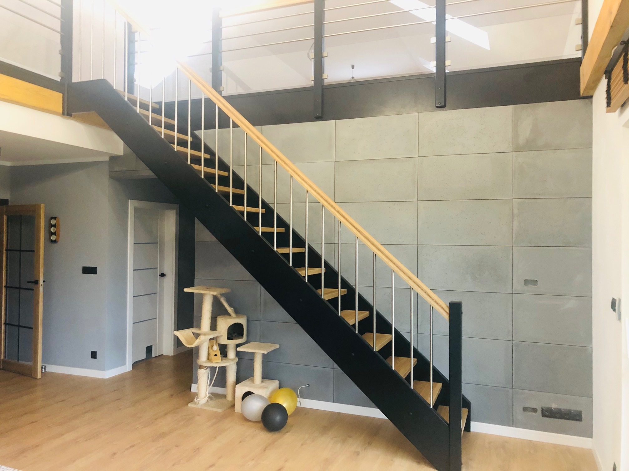 Nowoczesne schody z czarną konstrukcją, jasnymi drewnianymi stopniami i metalowymi balustradami w przestronnym wnętrzu z betonową ścianą i drewnianą podłogą, obok drapak dla kota i balony.