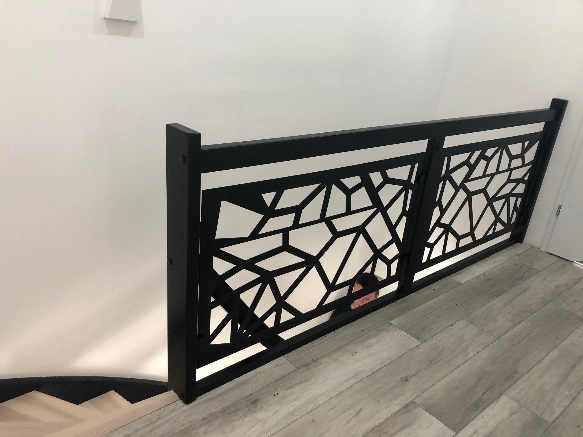 Nowoczesna czarna balustrada z geometrycznym ażurem, widok z góry na schody z drewnopodobnymi panelami, fragment osoby w tle.