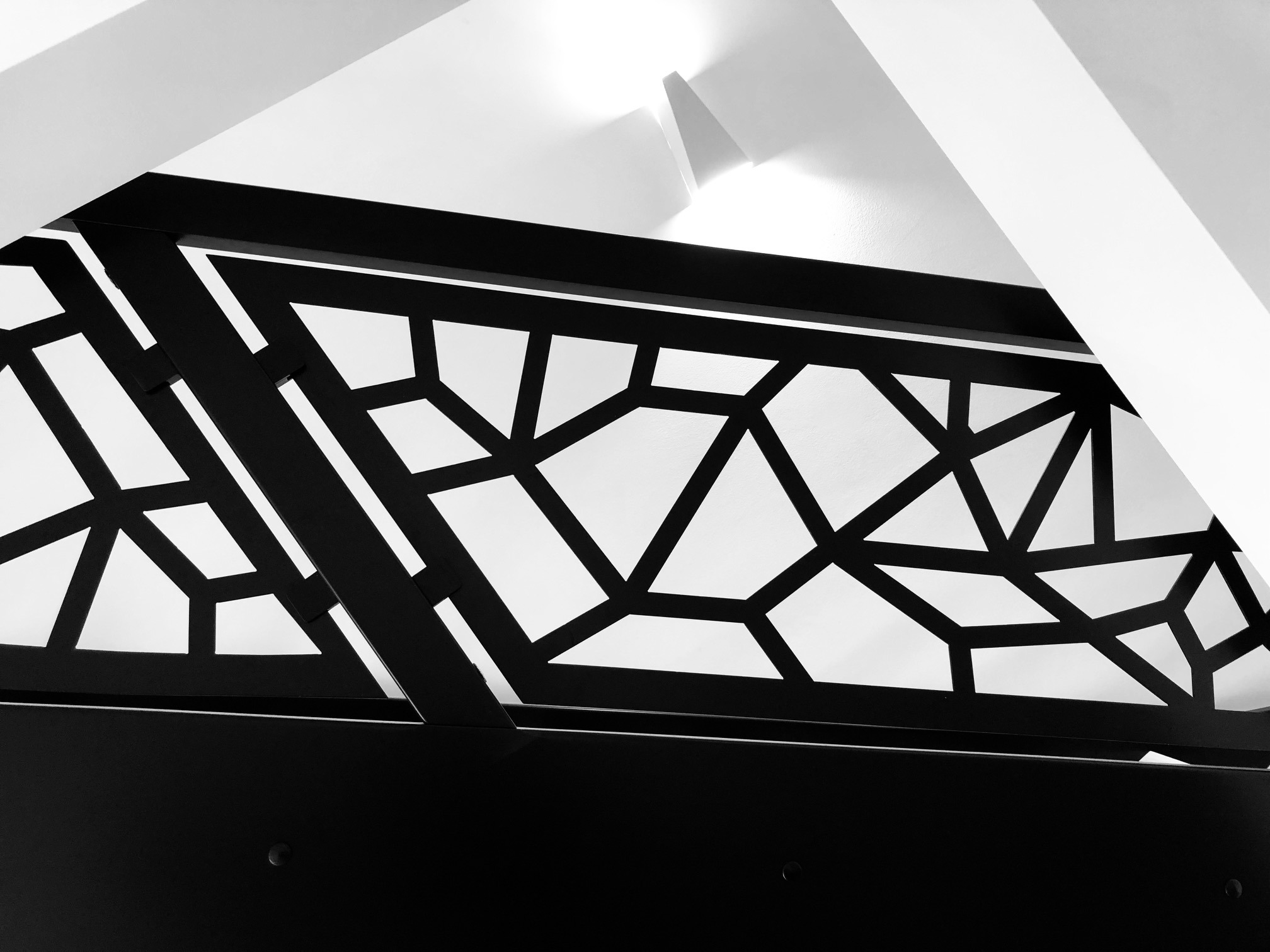 Czarno-białe ujęcie geometrycznego wzoru balustrady, z widocznymi elementami konstrukcyjnymi i fragmentem sufitu w tle.