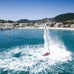 API Studio - Flyboard w akcji na tle portu jachtowego i plaży z zabudową miejską oraz górą w tle, woda wzburzona.