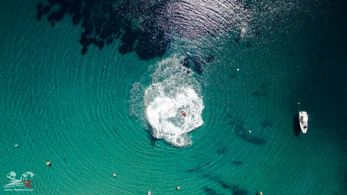 Widok z drona na osobę uprawiającą flyboard na turkusowej wodzie, z fontanną wody i łodzią w pobliżu.