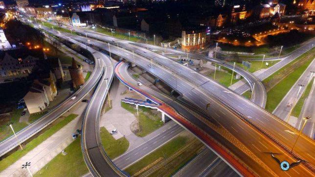 Nocna fotografia z lotu ptaka na wielopoziomowy węzeł drogowy z oświetlonymi budynkami w tle, wykonana z użyciem drona z widocznym obiektywem kamery.