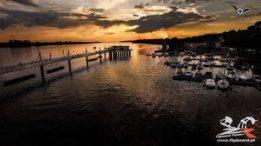 Panoramiczny widok z drona na przystań jachtową o zmierzchu, długi pomost z altaną, ciemne chmury na pomarańczowym niebie, logo firmy w prawym dolnym rogu.