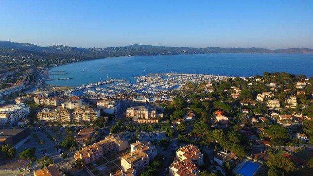 Panoramiczny widok z lotu ptaka na miasto portowe z mariną pełną jachtów, otoczone wzgórzami i morzem, sfotografowane w ciepłym świetle poranka.