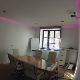 Jasne wnętrze sali konferencyjnej z dużym drewnianym stołem, krzesłami biurowymi, tablicą suchościeralną, oknem z żaluzjami i laptopem na stole, oświetlone fioletowymi lampami LED na suficie.