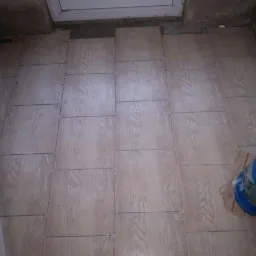 Układanie płytek podłogowych imitujących drewno, widoczne krzyżyki dystansowe, nieukończona praca, butla z gazem obok.