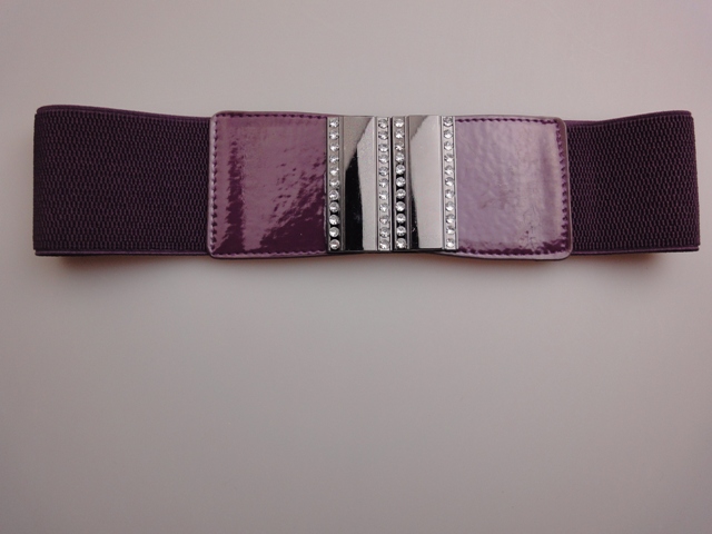 Elastyczny, fioletowy pasek damski z lakierowaną klamrą ozdobioną rzędami cyrkonii, leżący na gładkim, jasnym tle.