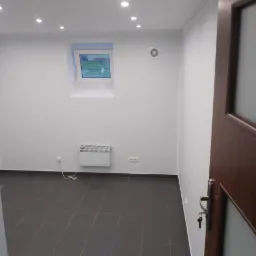 Wykończone wnętrze piwnicy zaadaptowanej na biuro, z ciemnymi płytkami na podłodze, białymi ścianami, grzejnikiem elektrycznym, małym oknem i oświetleniem punktowym w suficie, widoczne częściowo...