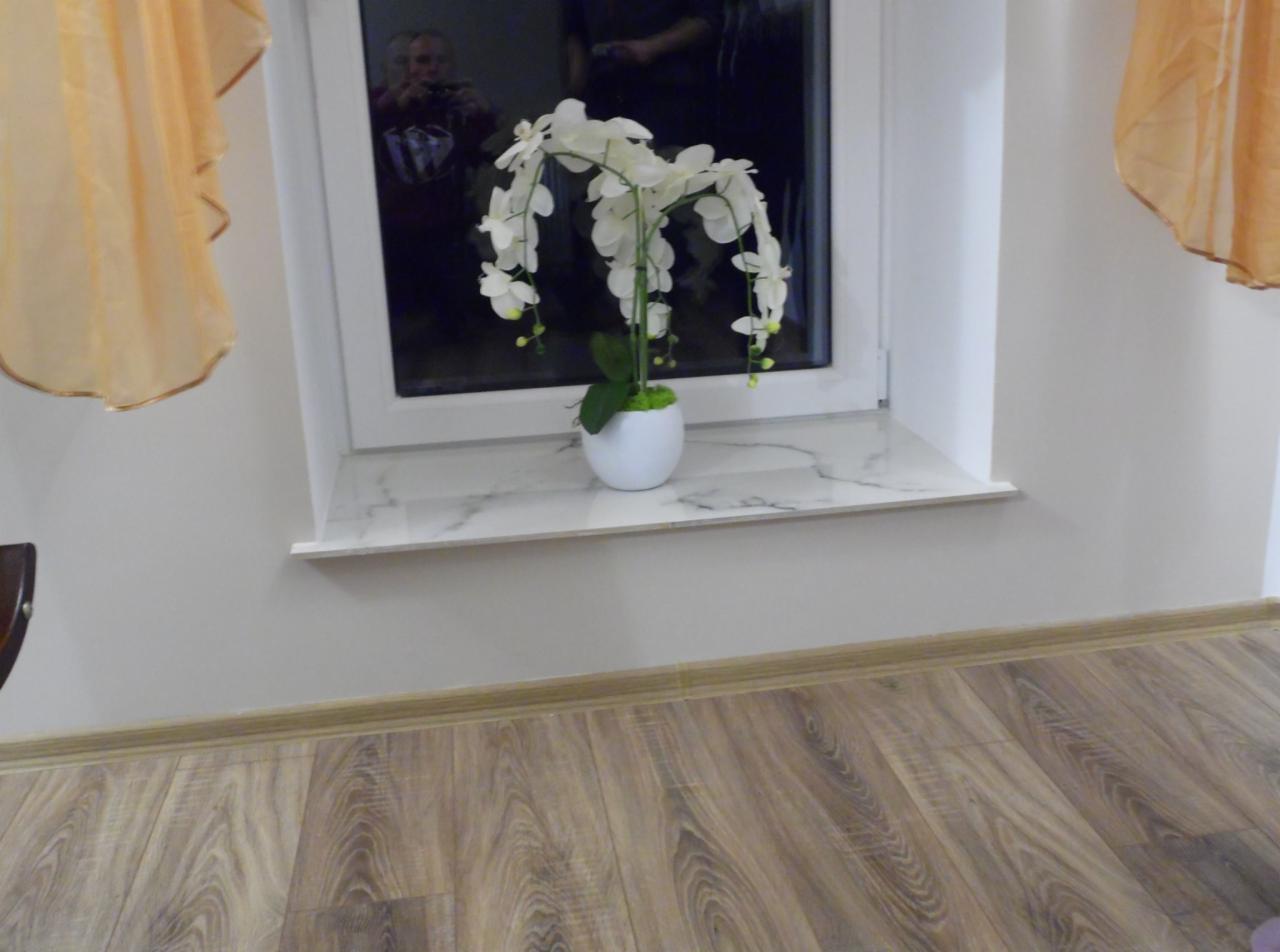 Biała orchidea w doniczce na marmurowym parapecie, widoczne panele podłogowe i fragment firanki, odbicie osoby w szybie okna.