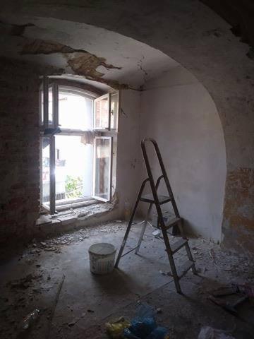 Pomieszczenie w trakcie adaptacji: widoczne fragmenty starego tynku na sklepieniu łukowym, okno z otwartymi skrzydłami, drabina przystawna, wiadro oraz gruz na podłodze.