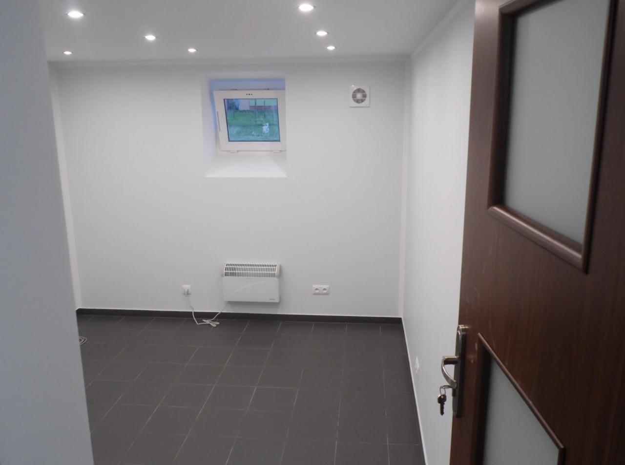 Wykończone wnętrze piwnicy zaadaptowanej na biuro, z ciemnymi płytkami na podłodze, białymi ścianami, grzejnikiem elektrycznym, małym oknem i oświetleniem punktowym w suficie, widoczne częściowo...