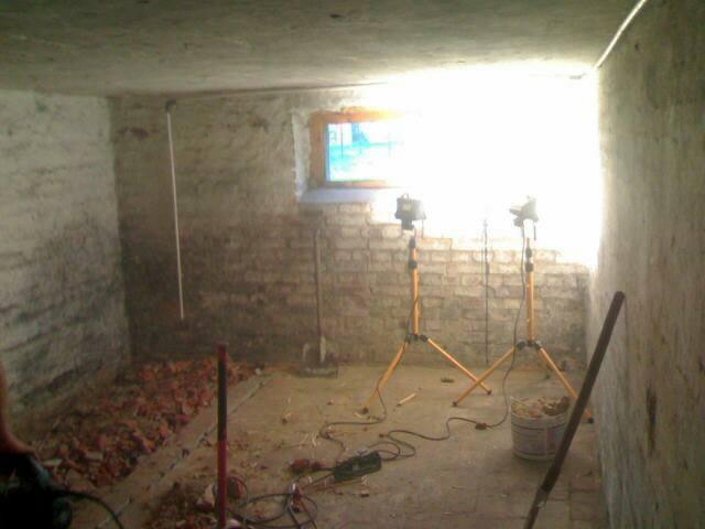 Piwnica w trakcie adaptacji na biuro: surowe ściany, gruz, dwa reflektory na trójnogach oświetlające pomieszczenie, widoczne okno i narzędzia pracy.