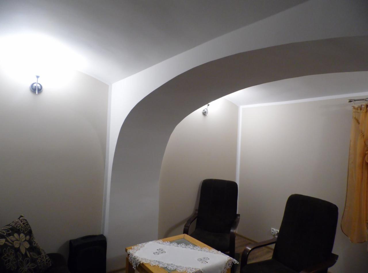 Adaptacja pomieszczenia gospodarczego: łukowate przejście między pokojami, ściany pomalowane na beżowo, widoczne dwa krzesła biurowe i stolik z serwetą.