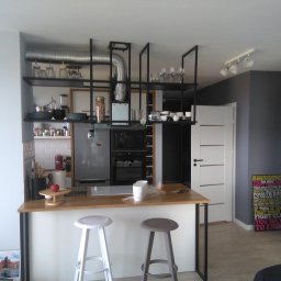 P.P.H.U. Meyer - Nowoczesna kuchnia w stylu industrialnym z wyspą, na której stoją dwa stołki barowe; nad wyspą zawieszona metalowa konstrukcja z półkami, na których znajdują się naczynia i akcesoria kuchenne.
