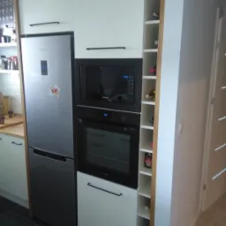 Nowoczesna zabudowa kuchenna z białymi frontami, czarnymi uchwytami, wbudowanym sprzętem AGD: lodówką, piekarnikiem i mikrofalówką Samsung oraz półkami na wino z boku.