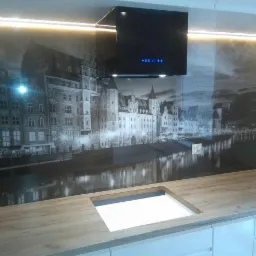 Nowoczesna kuchnia z blatem drewnianym o jasnym odcieniu, czarnym okapem i fototapetą przedstawiającą panoramę miasta nad rzeką w odcieniach szarości.