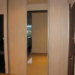 Trzydrzwiowa szafa przesuwna z lustrzanym środkowym panelem, jasne drewnopodobne fronty, widoczny fragment wnętrza mieszkania.