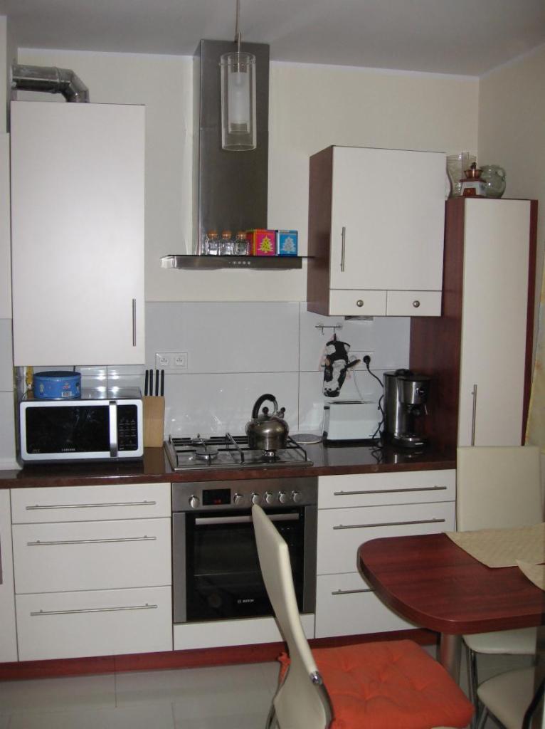 Wnętrze kuchni z białymi szafkami, brązowym blatem, kuchenką gazową i piekarnikiem, widoczny fragment stołu z krzesłami i pomarańczową poduszką.