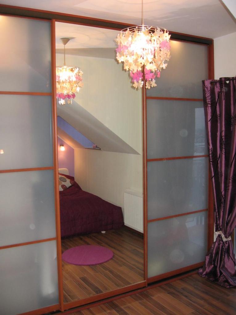 Szafa przesuwna z lustrzanymi drzwiami odbijająca wnętrze pokoju na poddaszu z fioletowym dywanikiem i łóżkiem, widoczna dekoracyjna lampa z motywem kwiatowym.