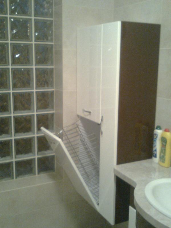 Szafka łazienkowa z otwieranym koszem na pranie, widoczna ściana z luksferami, jasne płytki i fragment umywalki z blatem.